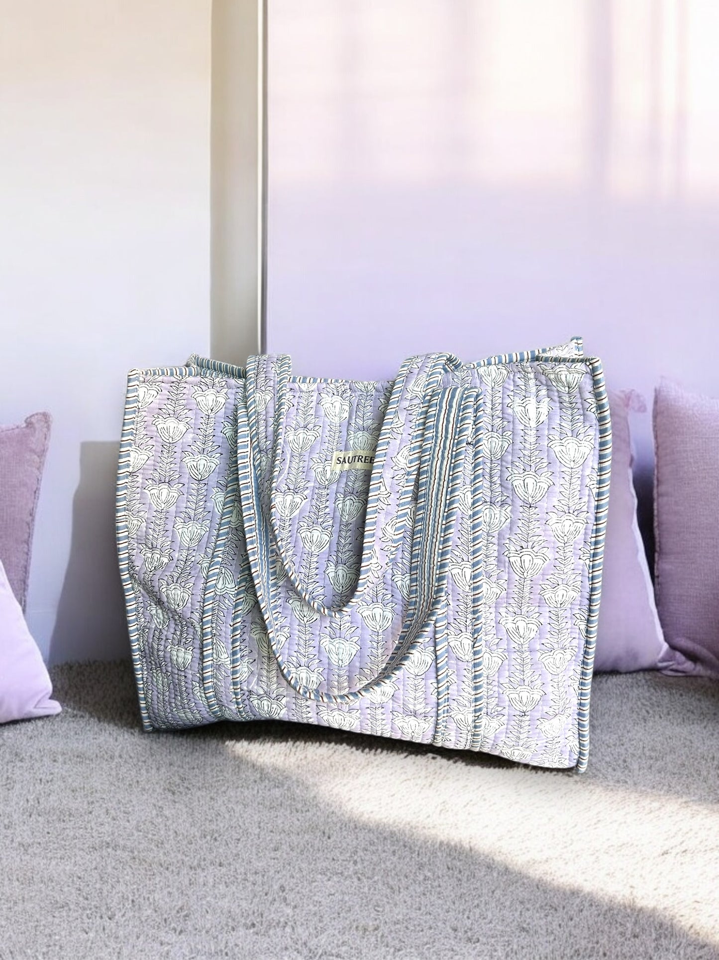 A hand block printed tote bag with a floral pattern in lilac and blue colors, displayed on a floor against a wall.